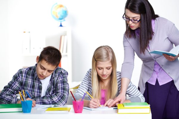 Portrait of teenage lad and girl writing somethinig during lesson while teacher checking their notes