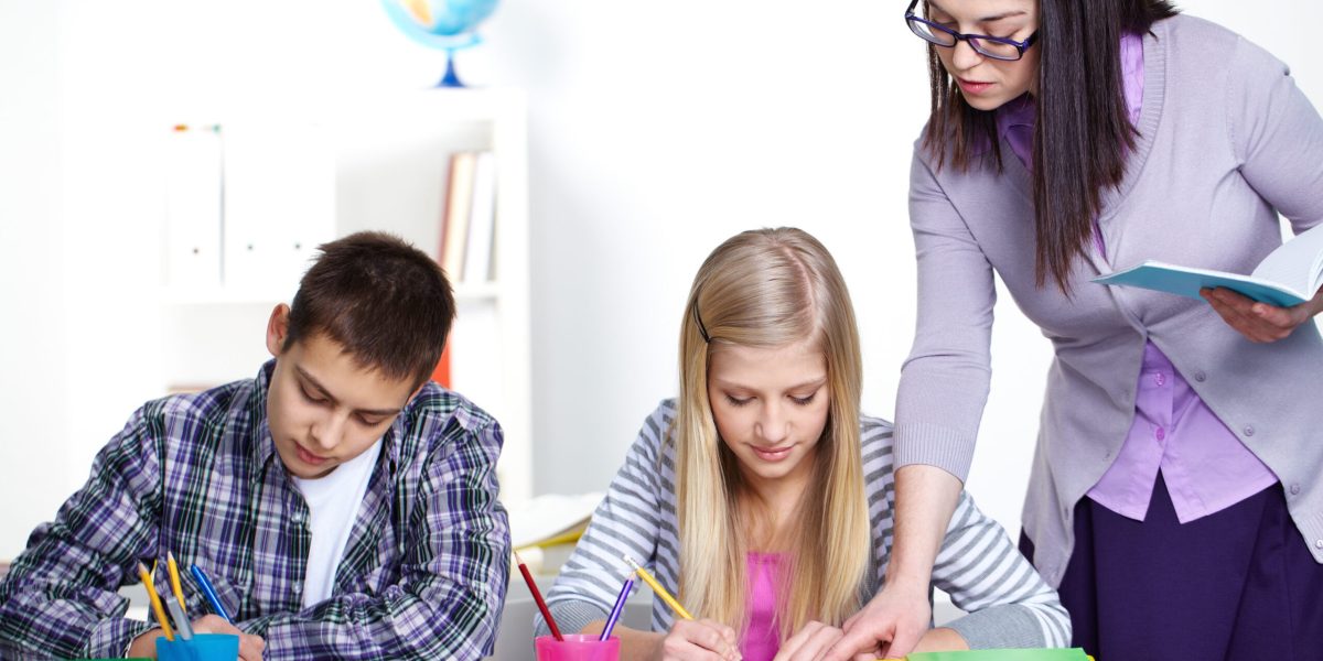 Portrait of teenage lad and girl writing somethinig during lesson while teacher checking their notes